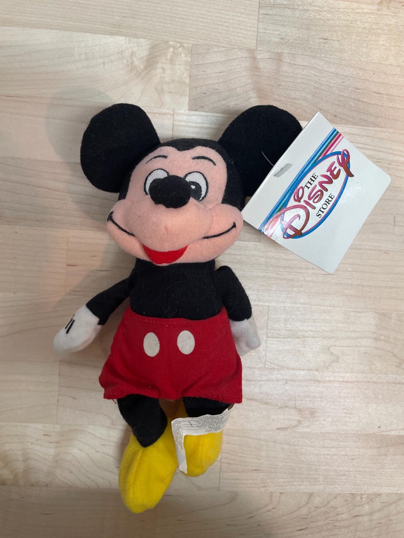 May include: A Mickey Mouse soft toy with black ears, a black body, red shorts with white buttons, yellow shoes, and white gloves. The toy has a tag that says "The Disney Store".