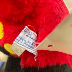 May include: Close-up of a red and black plush Mickey Mouse toy with a white tag that reads "House of Taiwan, Fiber, Foam, Disney Prods."