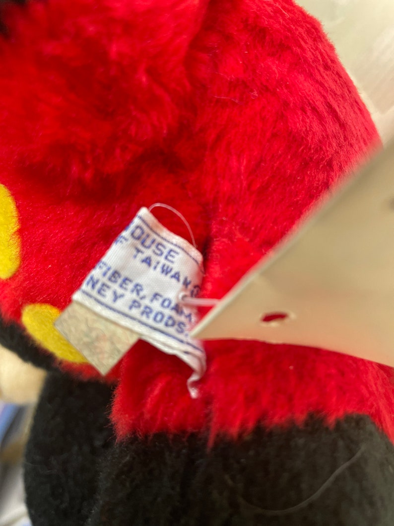 May include: Close-up of a red and black plush Mickey Mouse toy with a white tag that reads "House of Taiwan, Fiber, Foam, Disney Prods."