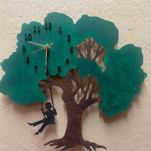 May include: A wall clock shaped like a tree with a green canopy and a brown trunk. A silhouette of a person on a swing hangs from a branch. The clock face features black numbers from 1 to 12 and gold-colored hour, minute, and second hands.