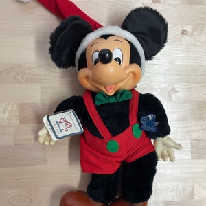 May include: A plush Mickey Mouse wearing a red and white Santa hat and red overalls with green accents. The plush toy is holding a tag that says "Applause" and "To Beat of Music".