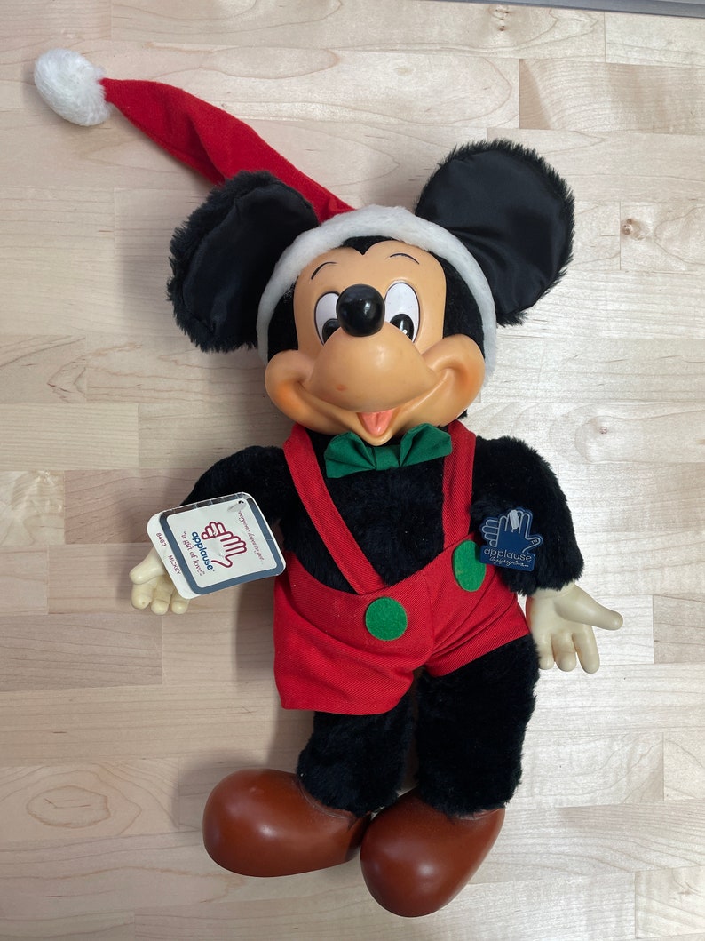 May include: A plush Mickey Mouse wearing a red and white Santa hat and red overalls with green accents. The plush toy is holding a tag that says "Applause" and "To Beat of Music".