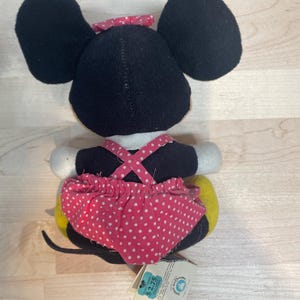 Puede incluir: Un peluche de Minnie Mouse, visto desde atr&aacute;s, presenta grandes orejas negras y un lazo rojo con lunares. El juguete lleva una falda rosa con lunares y una parte superior negra con tirantes rosas. Se ve una cola amarilla y una etiqueta.