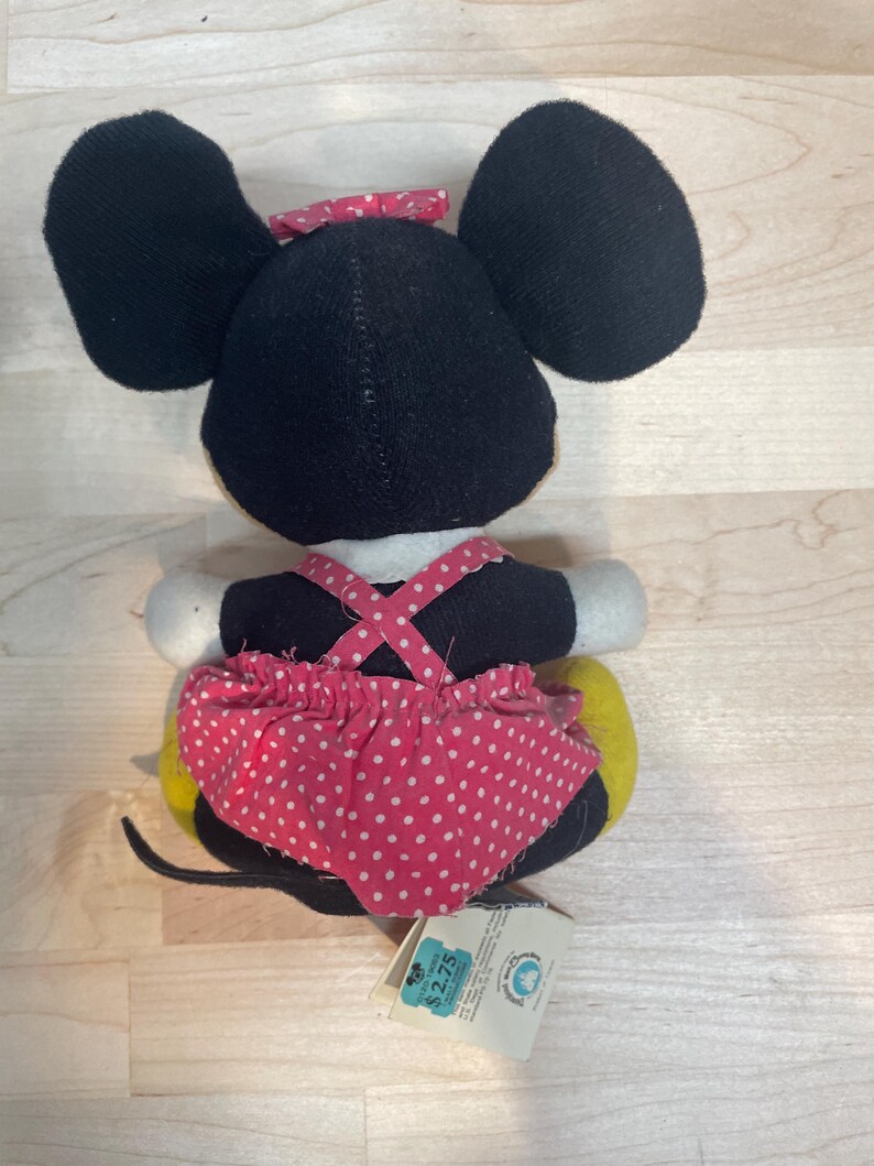 Puede incluir: Un peluche de Minnie Mouse, visto desde atr&aacute;s, presenta grandes orejas negras y un lazo rojo con lunares. El juguete lleva una falda rosa con lunares y una parte superior negra con tirantes rosas. Se ve una cola amarilla y una etiqueta.