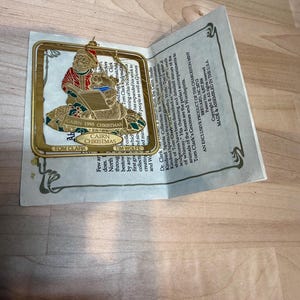 May include: A gold-colored Christmas ornament featuring a seated Santa Claus reading a book, with the text "CAIRN CHRISTMAS 1995". The ornament is in a rectangular frame with a decorative border. The ornament is on a printed card.