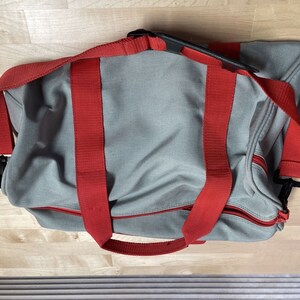 Puede incluir: Una bolsa de lona gris con correas rojas y tiradores de cremallera negros.