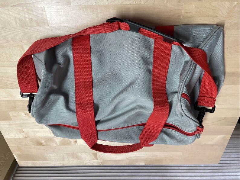 Puede incluir: Una bolsa de lona gris con correas rojas y tiradores de cremallera negros.