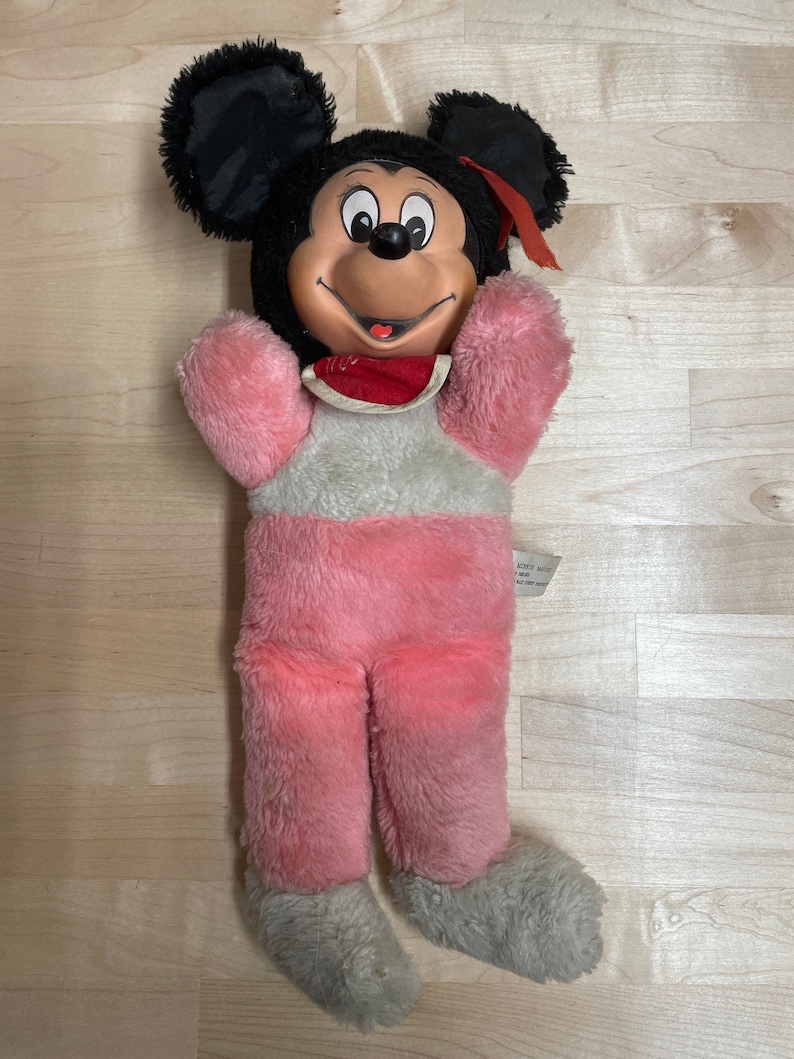 Peut inclure: Un jouet en peluche Mickey Mouse rose et gris avec des oreilles noires et un n&oelig;ud rouge. Le jouet a un visage souriant avec des yeux noirs et une bouche rouge.