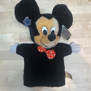 May include: A plush Mickey Mouse hand puppet with a black body, large black ears, and a smiling face. The puppet has a red bow tie with white polka dots and gray hands. Tags are attached to the puppet.