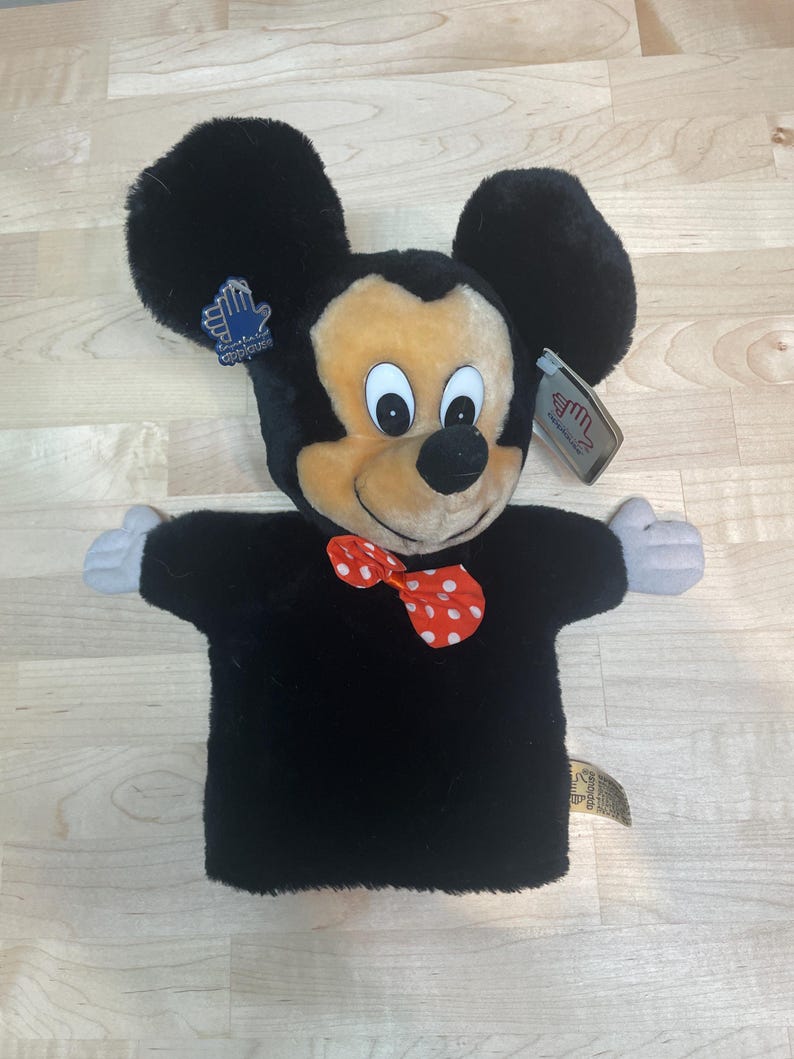 May include: A plush Mickey Mouse hand puppet with a black body, large black ears, and a smiling face. The puppet has a red bow tie with white polka dots and gray hands. Tags are attached to the puppet.