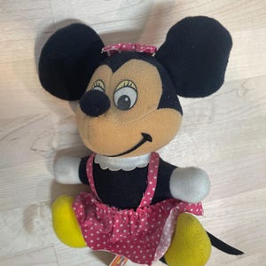 Puede incluir: Un peluche vintage de Minnie Mouse. El peluche presenta orejas negras, una cara color canela y una falda rosa con lunares. El juguete tiene pies amarillos, guantes blancos y un peque&ntilde;o lazo rosa en la cabeza. Una peque&ntilde;a etiqueta est&aacute; adjunta.