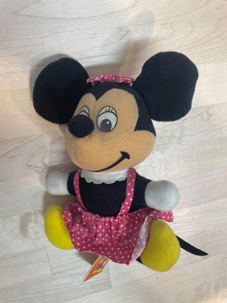 Puede incluir: Un peluche vintage de Minnie Mouse. El peluche presenta orejas negras, una cara color canela y una falda rosa con lunares. El juguete tiene pies amarillos, guantes blancos y un peque&ntilde;o lazo rosa en la cabeza. Una peque&ntilde;a etiqueta est&aacute; adjunta.