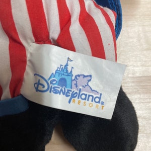 K&ouml;nnte beinhalten: Schwarz-wei&szlig;es Pl&uuml;schtier mit rot-wei&szlig; gestreiftem Stoff. Das Spielzeug hat einen blauen Kragen und ein Disneyland Resort-Etikett.