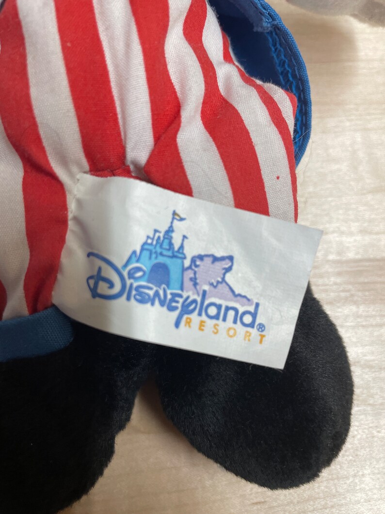 K&ouml;nnte beinhalten: Schwarz-wei&szlig;es Pl&uuml;schtier mit rot-wei&szlig; gestreiftem Stoff. Das Spielzeug hat einen blauen Kragen und ein Disneyland Resort-Etikett.