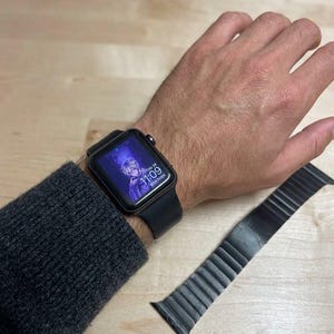 May include: A black smartwatch with a rectangular screen displaying an anime-style character, the time 11:09, and the date Thu 28. The watch features a smooth black silicone band, and a separate black magnetic leather link band is placed nearby.