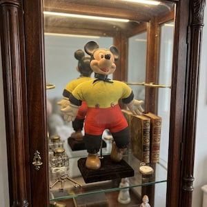 May include: A vintage Mickey Mouse doll featuring a yellow shirt with black cuffs, red shorts with two buttons, and brown shoes. The figure has a black neck ribbon, white gloves, and a classic molded face, standing on a dark square base.