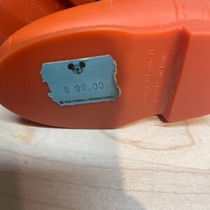 Pu&ograve; includere: Primo piano di uno stivale vintage in plastica arancione con un polsino in denim blu. Lo stivale ha un'etichetta del prezzo azzurra con la scritta "$ 20.00" e un logo di Topolino. Il testo "YOUNG EPOCH MADE IN JAPAN" &egrave; visibile.