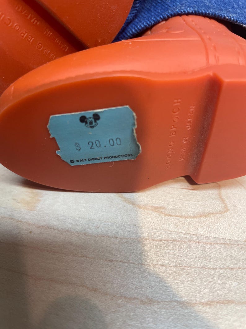 Pu&ograve; includere: Primo piano di uno stivale vintage in plastica arancione con un polsino in denim blu. Lo stivale ha un'etichetta del prezzo azzurra con la scritta "$ 20.00" e un logo di Topolino. Il testo "YOUNG EPOCH MADE IN JAPAN" &egrave; visibile.