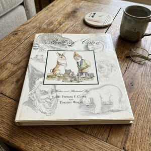 May include: A hardcover book titled Book of Cairn written and illustrated by Dr. Thomas F. Clark and Timothy Wolfe. The white cover features a central color illustration of a rabbit and a gnome, surrounded by various black and white sketches.