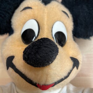 May include: Close-up of a plush Mickey Mouse toy with black ears, a black nose, and white eyes. The toy is wearing a white shirt.