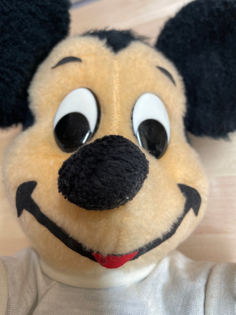 May include: Close-up of a plush Mickey Mouse toy with black ears, a black nose, and white eyes. The toy is wearing a white shirt.
