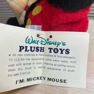May include: A white tag with black text reads "Walt Disney's Plush Toys" and includes instructions for cleaning the plush toy. The tag is attached to a red and black plush Mickey Mouse toy. The tag states "All new materials, Hand-crafted, Non-allergenic. To Clean-We recommend using warm water, mild soap with sponge or cloth, cleaning as soiling occurs. When dry, brush to restore appearance of plush. For ages 3 and up. I'm: Mickey Mouse."