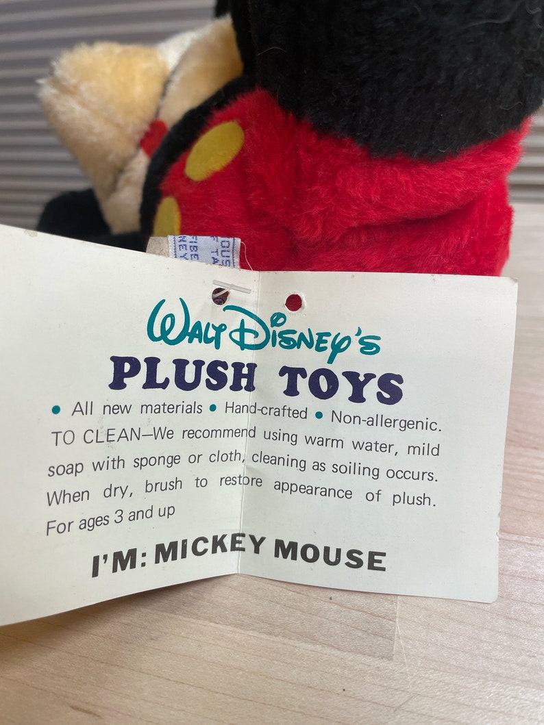 May include: A white tag with black text reads "Walt Disney's Plush Toys" and includes instructions for cleaning the plush toy. The tag is attached to a red and black plush Mickey Mouse toy. The tag states "All new materials, Hand-crafted, Non-allergenic. To Clean-We recommend using warm water, mild soap with sponge or cloth, cleaning as soiling occurs. When dry, brush to restore appearance of plush. For ages 3 and up. I'm: Mickey Mouse."