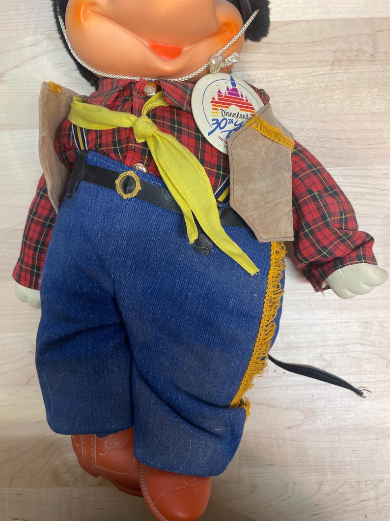 Pu&ograve; includere: Bambola vintage di Topolino con camicia a quadri rossa, jeans blu e gilet marrone. La bambola ha un cravattino giallo e un'etichetta del 30&deg; anniversario di Disneyland. La bambola indossa stivali arancioni.