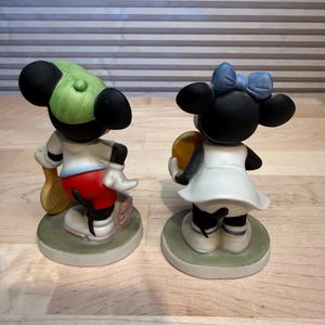 Puede incluir: Dos figuras de cer&aacute;mica de personajes de dibujos animados. Uno lleva una gorra verde, pantalones cortos rojos y sostiene una raqueta de tenis. El otro lleva un vestido blanco y un lazo azul, sosteniendo una pelota amarilla. Ambos est&aacute;n sobre bases redondas verdes.