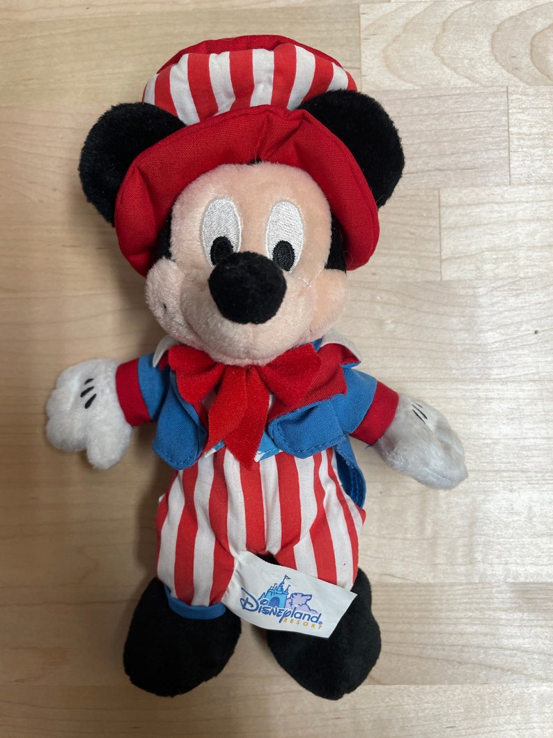 K&ouml;nnte beinhalten: Eine rot-wei&szlig;-blaue Pl&uuml;sch-Mickey-Mouse-Puppe mit einem rot-wei&szlig; gestreiften Hut und einer blauen Jacke mit einer roten Schleife. Die Puppe tr&auml;gt eine rot-wei&szlig; gestreifte Hose und hat ein schwarzes Disneyland Resort-Etikett an ihrem Fu&szlig;.