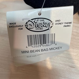 May include: A white tag featuring the Disney Store logo and the text "MADE EXCLUSIVELY FOR" and "MINI BEAN BAG MICKEY". The tag is attached to a black plush toy.