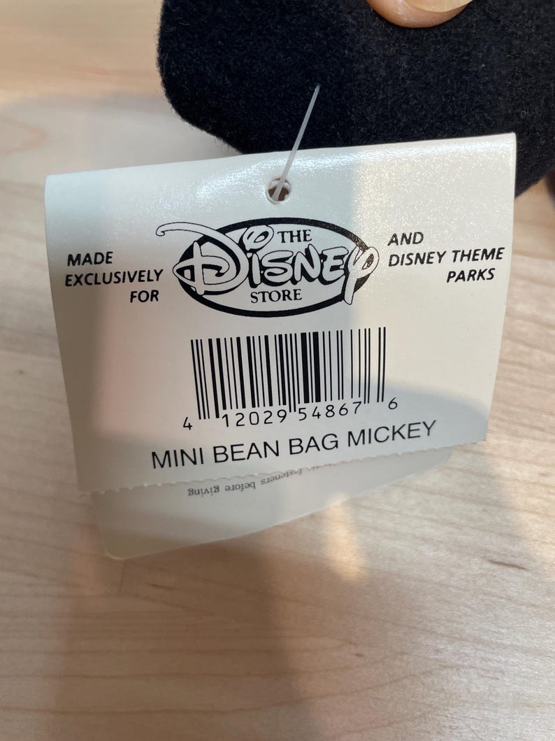 May include: A white tag featuring the Disney Store logo and the text "MADE EXCLUSIVELY FOR" and "MINI BEAN BAG MICKEY". The tag is attached to a black plush toy.