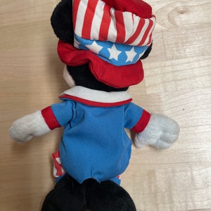 K&ouml;nnte beinhalten: Eine Pl&uuml;sch-Mickey-Mouse-Puppe, die einen rot-wei&szlig;-blauen Hut und ein Outfit tr&auml;gt. Der Hut hat einen roten Rand und eine blaue Oberseite mit wei&szlig;en Sternen. Das Outfit ist ein blaues Hemd mit rotem Kragen und wei&szlig;en Streifen an den &Auml;rmeln und der Hose.