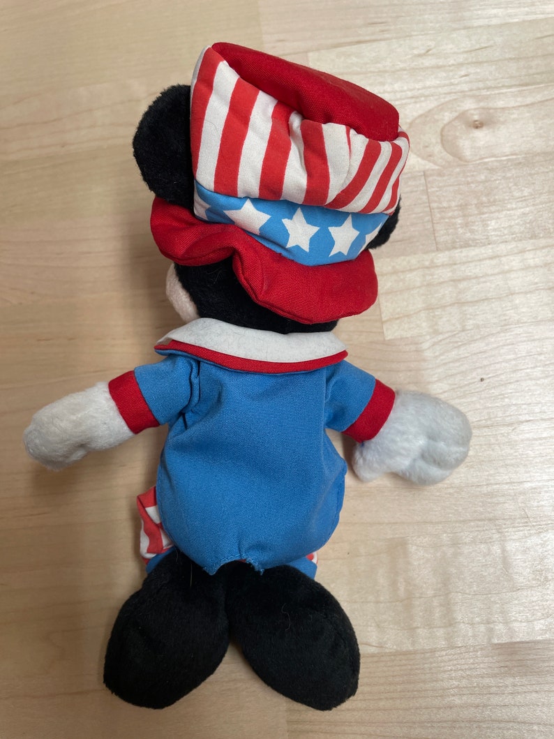 K&ouml;nnte beinhalten: Eine Pl&uuml;sch-Mickey-Mouse-Puppe, die einen rot-wei&szlig;-blauen Hut und ein Outfit tr&auml;gt. Der Hut hat einen roten Rand und eine blaue Oberseite mit wei&szlig;en Sternen. Das Outfit ist ein blaues Hemd mit rotem Kragen und wei&szlig;en Streifen an den &Auml;rmeln und der Hose.