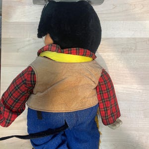 Pu&ograve; includere: Una bambola cowboy vintage, vista da dietro, con un cappello nero, una camicia a quadri rossi, un gilet marrone e pantaloni di jeans blu. La bambola ha un colletto giallo e stivali arancioni. Il cappello della bambola &egrave; attaccato con una catena d'argento.