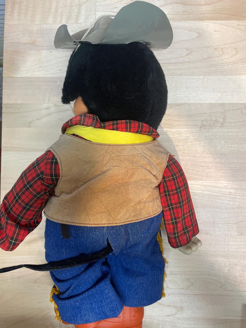 Pu&ograve; includere: Una bambola cowboy vintage, vista da dietro, con un cappello nero, una camicia a quadri rossi, un gilet marrone e pantaloni di jeans blu. La bambola ha un colletto giallo e stivali arancioni. Il cappello della bambola &egrave; attaccato con una catena d'argento.