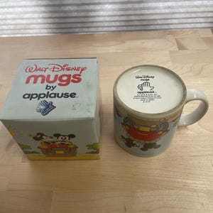 K&ouml;nnte beinhalten: Ein Vintage-Set aus Walt Disney "Mugs by applause" Tasse und Box. Die Tasse zeigt ein Cartoon-Design mit Micky Maus und anderen Figuren. Die Box ist hellblau mit den Worten "Walt Disney mugs by applause" in Rot.