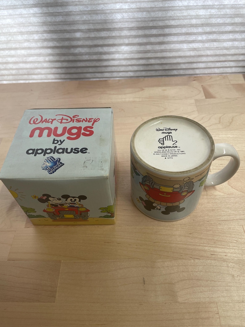 K&ouml;nnte beinhalten: Ein Vintage-Set aus Walt Disney "Mugs by applause" Tasse und Box. Die Tasse zeigt ein Cartoon-Design mit Micky Maus und anderen Figuren. Die Box ist hellblau mit den Worten "Walt Disney mugs by applause" in Rot.