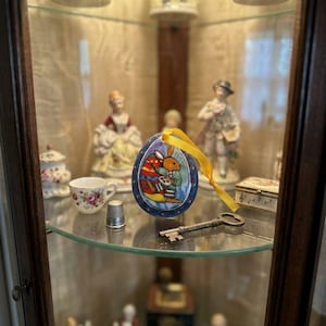 Könnte beinhalten: Ein eiförmiges Keramikornament mit einem bemalten Hasen, der eine Malerpalette hält, verziert mit einem blauen Rand und einer gelben Schleife. Das Ornament steht auf einem Glasregal neben einem Metallfingerhut und einem alten Metallschlüssel.