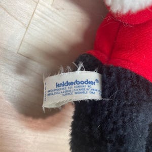 Puede incluir: Un peluche con un abrigo rojo y adornos blancos. El juguete tiene pelo negro y una etiqueta que dice "knickerbocker". La etiqueta tambi&eacute;n incluye la direcci&oacute;n de la empresa y la frase "surface washable only."