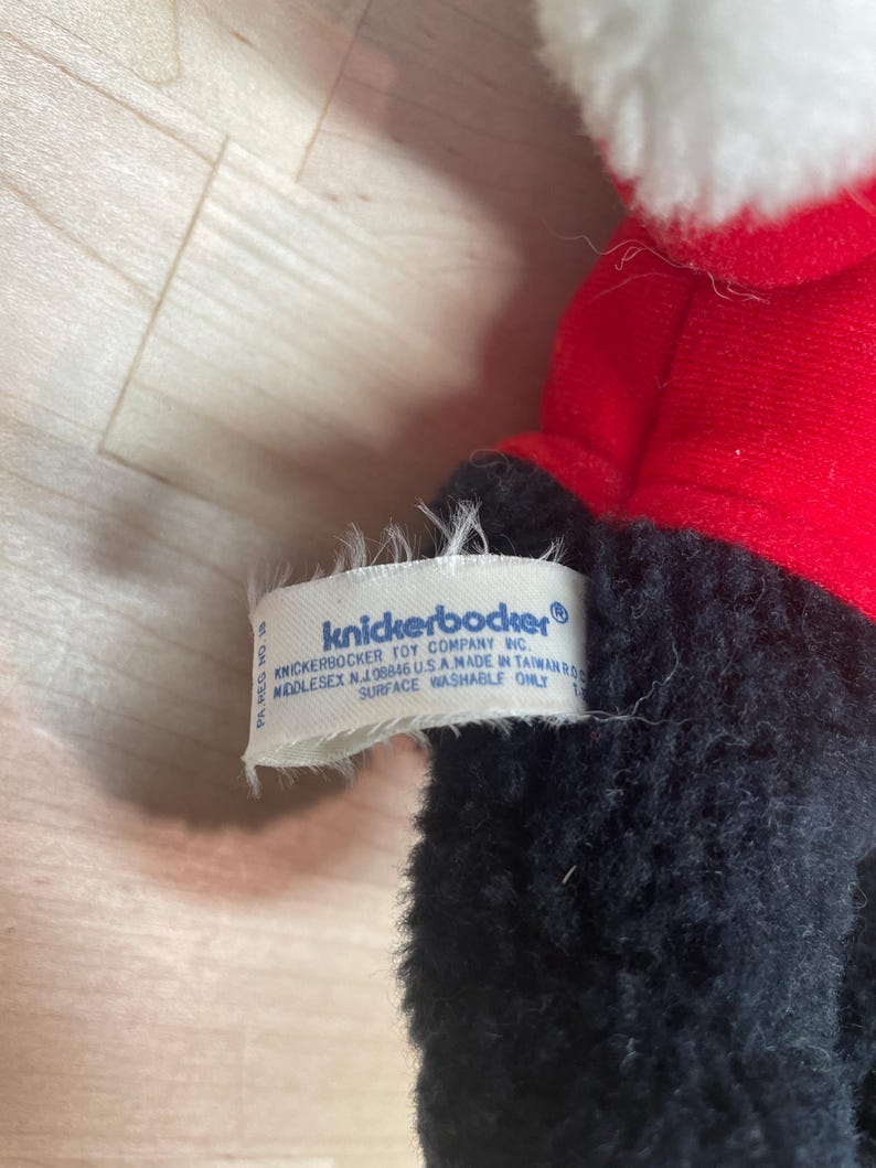 Puede incluir: Un peluche con un abrigo rojo y adornos blancos. El juguete tiene pelo negro y una etiqueta que dice "knickerbocker". La etiqueta tambi&eacute;n incluye la direcci&oacute;n de la empresa y la frase "surface washable only."