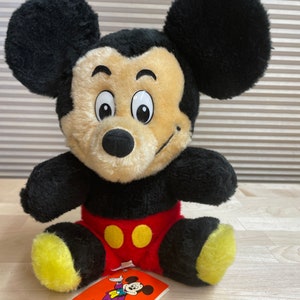 May include: A plush black and yellow Mickey Mouse toy sitting on a wooden surface. The toy is wearing red shorts with yellow buttons. A small tag with a cartoon image of Mickey Mouse is in front of the toy.