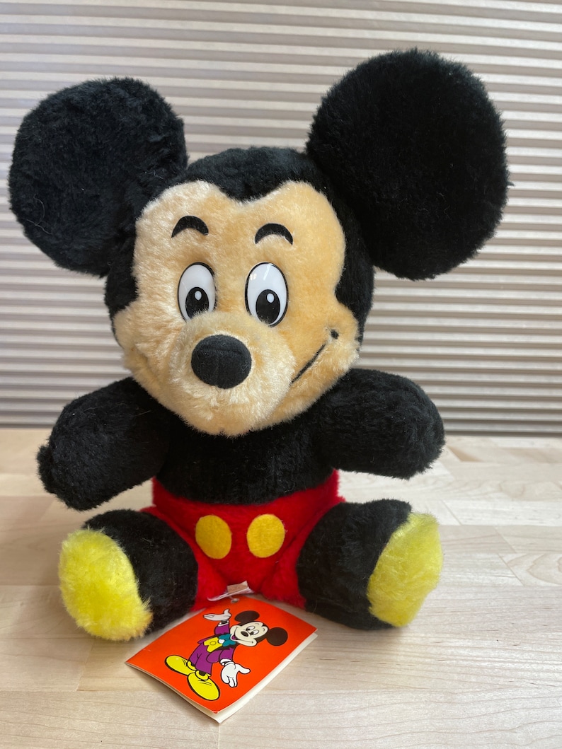 May include: A plush black and yellow Mickey Mouse toy sitting on a wooden surface. The toy is wearing red shorts with yellow buttons. A small tag with a cartoon image of Mickey Mouse is in front of the toy.