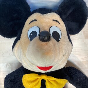 K&ouml;nnte beinhalten: Eine Mickey Mouse-Pl&uuml;schpuppe mit gro&szlig;en schwarzen Ohren, einem beigefarbenen Gesicht und einem roten L&auml;cheln. Er tr&auml;gt einen schwarzen K&ouml;rper, eine gelbe Fliege und rote Shorts. Die Puppe hat gro&szlig;e, wei&szlig; umrandete Augen und eine schwarze Nase.