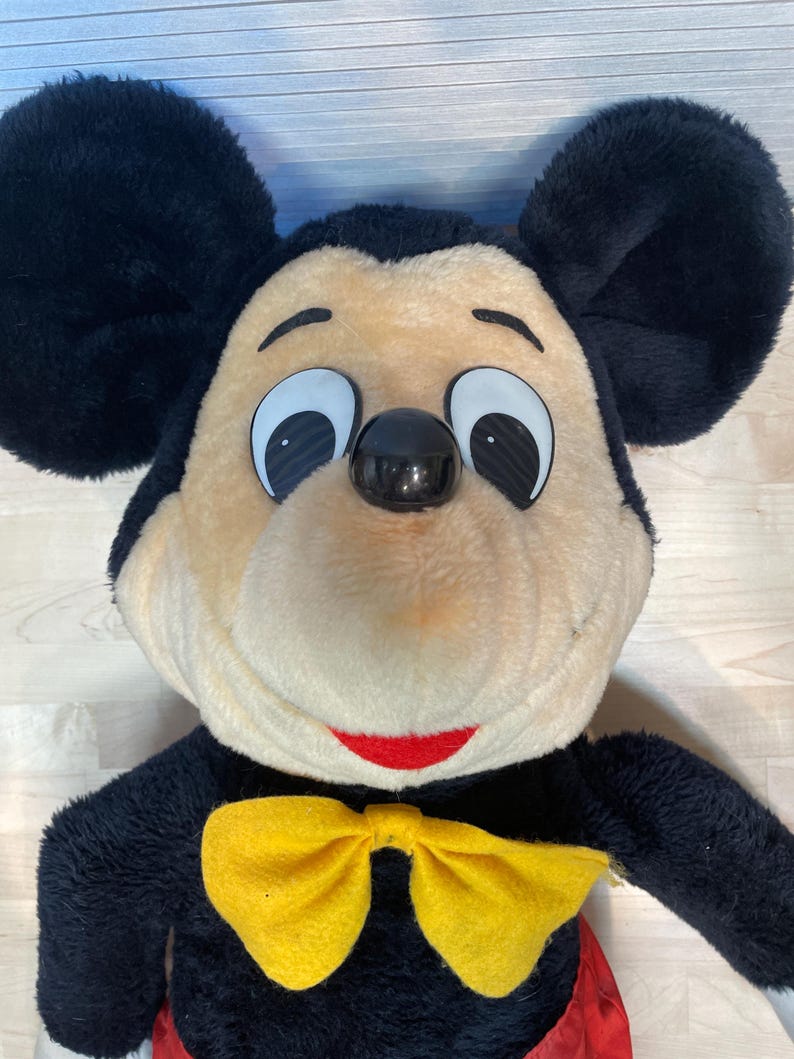 K&ouml;nnte beinhalten: Eine Mickey Mouse-Pl&uuml;schpuppe mit gro&szlig;en schwarzen Ohren, einem beigefarbenen Gesicht und einem roten L&auml;cheln. Er tr&auml;gt einen schwarzen K&ouml;rper, eine gelbe Fliege und rote Shorts. Die Puppe hat gro&szlig;e, wei&szlig; umrandete Augen und eine schwarze Nase.