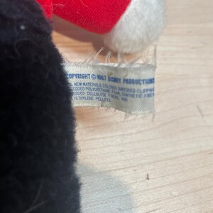 Puede incluir: Un peluche con cuerpo negro, gorro rojo y ribetes blancos. Una etiqueta dice "COPYRIGHT &copy; WALT DISNEY PRODUCTIONS" y enumera los materiales. Probablemente un objeto de colecci&oacute;n vintage.
