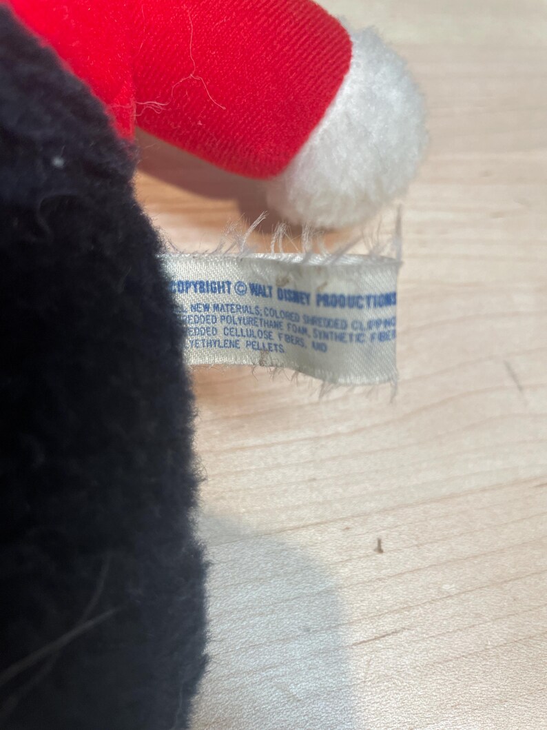 Puede incluir: Un peluche con cuerpo negro, gorro rojo y ribetes blancos. Una etiqueta dice "COPYRIGHT &copy; WALT DISNEY PRODUCTIONS" y enumera los materiales. Probablemente un objeto de colecci&oacute;n vintage.