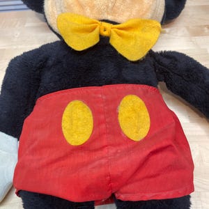 K&ouml;nnte beinhalten: Eine Mickey Mouse Pl&uuml;schpuppe mit schwarzem Fell, einem beigen Gesicht und einer gelben Fliege. Die Puppe tr&auml;gt rote Shorts mit zwei gelben Kreisen. Die Puppenmerkmale umfassen schwarze Ohren, einen roten Mund und wei&szlig;e Handschuhe.