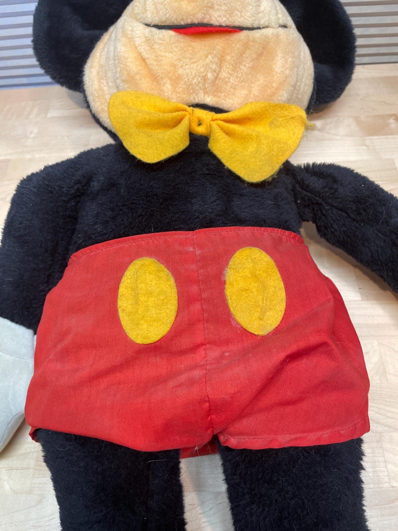 K&ouml;nnte beinhalten: Eine Mickey Mouse Pl&uuml;schpuppe mit schwarzem Fell, einem beigen Gesicht und einer gelben Fliege. Die Puppe tr&auml;gt rote Shorts mit zwei gelben Kreisen. Die Puppenmerkmale umfassen schwarze Ohren, einen roten Mund und wei&szlig;e Handschuhe.