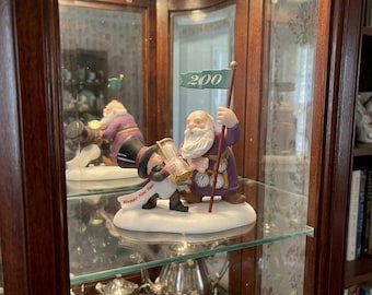 May include: A ceramic figurine set featuring a bearded figure in a purple robe with clock motifs and a smaller figure in a top hat holding an hourglass. The larger figure holds a green flag with the text 200. Both stand on a white base.